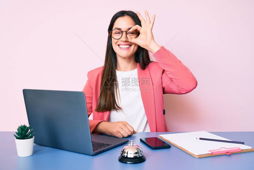 坐在接待员办公桌前工作的年轻白人女用笔记本电脑微笑着开心地做ok手势图片