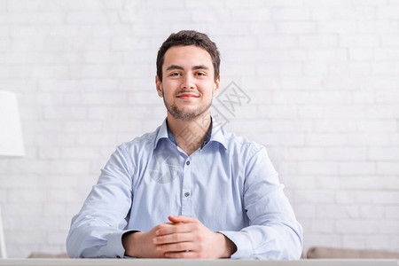 在家进行视频会议身穿衬衫的正式微笑男坐在白砖墙背景的桌子旁图片