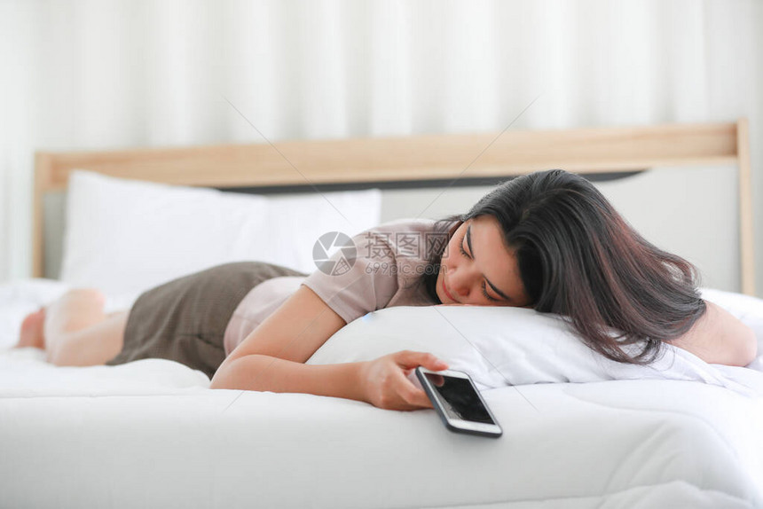 美丽的亚洲女人躺在卧室的床上她手里拿着智能手机睡觉年轻女现代生活方式的互联网社交和电图片