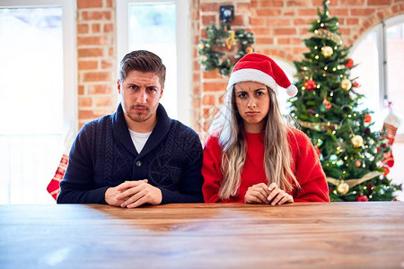一对戴着圣诞老人帽的年轻夫妇坐在家里圣诞树周围的椅子和桌子上图片