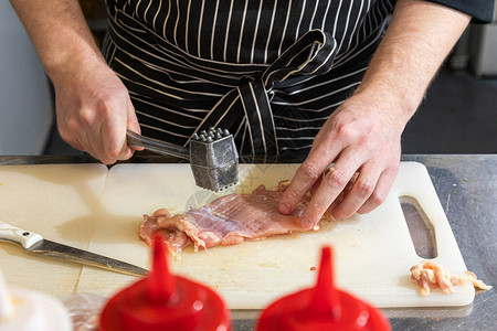 穿黑围裙的近身男主厨在专业厨房用铁锤砸了一块鸡肉图片