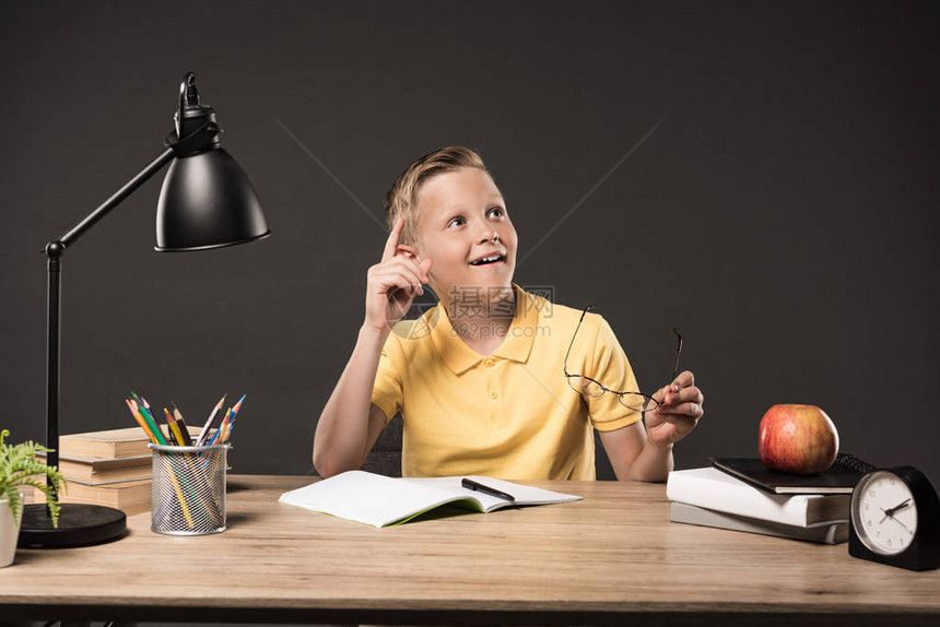 男孩在餐桌上用灰色背景的书植物灯彩铅笔苹果钟表和教科书手持眼镜图片