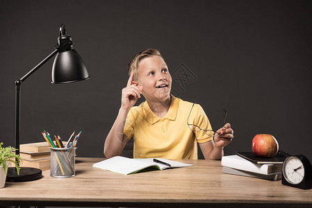男孩在餐桌上用灰色背景的书植物灯彩铅笔苹果钟表和教科书手持眼镜图片