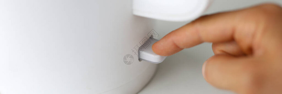零度下沸腾用手指按下电水壶上的旋钮的人特写镜头打开电源温水泡茶或咖啡现代设备桌上的白色茶壶背景
