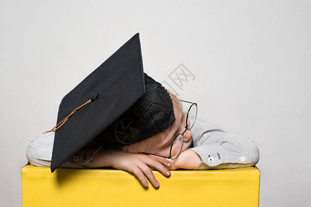 戴着学术帽子和戴眼镜睡在桌子上的小金发男孩图片