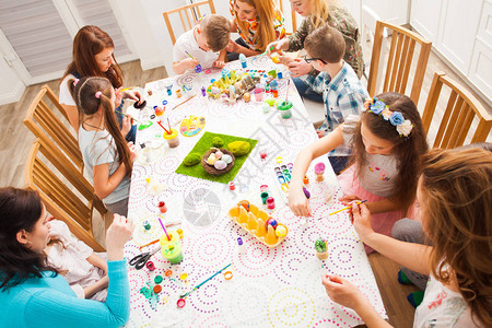 儿童坐在大桌旁装饰和绘画传统复活节鸡蛋的首版照片成年妇女帮助儿童创意讲习背景图片