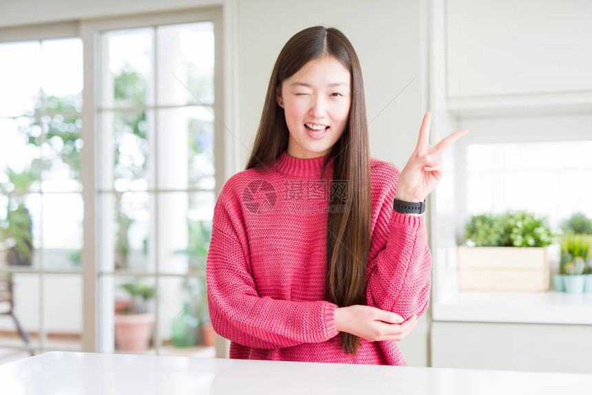 美丽的亚洲女人在白桌上穿着粉红色毛衣笑着面朝镜头眨眼看拍图片