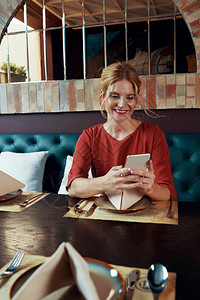 坐在餐桌发短信或使用移动应用程序的穿着亮红礼服的美丽笑着微笑图片
