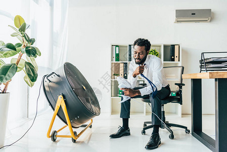 AfricanAmerican商人坐在办公室的电通风机前图片