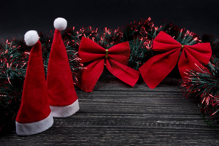 两顶圣诞帽两顶红色圣诞蝴蝶结和黑色木质背景上的绿色和红色金属箔图片