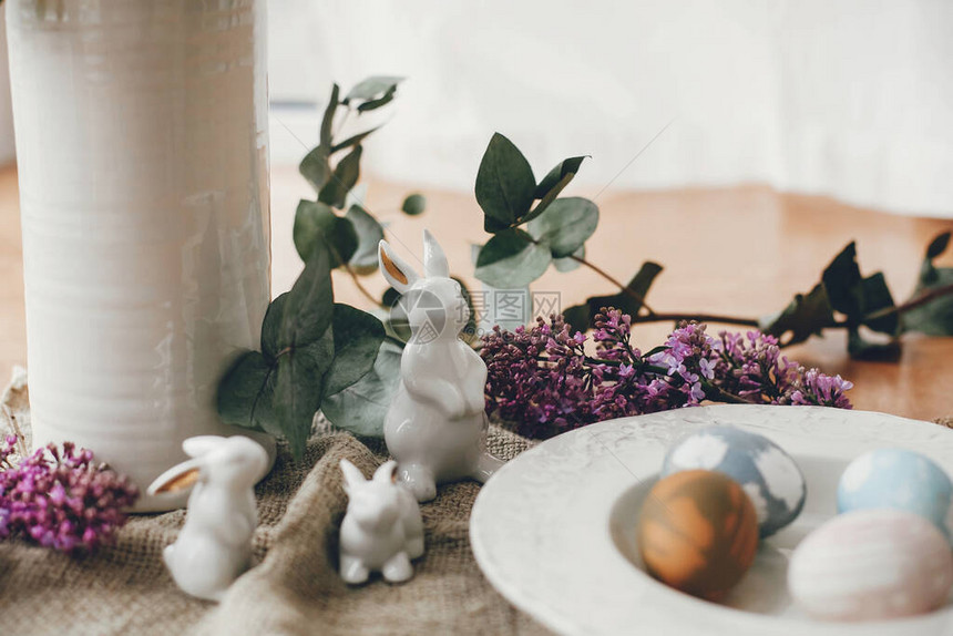 复古盘子上的时尚复活节彩蛋白色小兔子和木桌上质朴织物上的丁香花天然染色鸡蛋和春紫花的农村图片