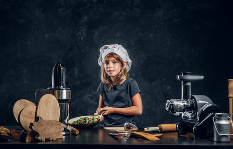 戴厨师帽的可爱小女孩正准备蔬菜在黑暗背景下做饭图片