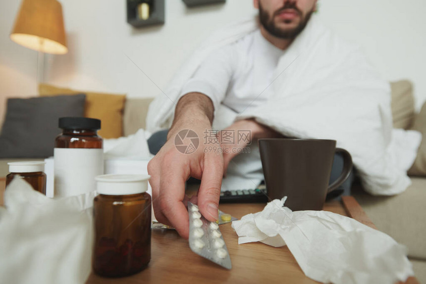 生病的年轻男子在家中单独与自己隔绝时坐在床上或沙发上时用止痛药或预产物从桌图片