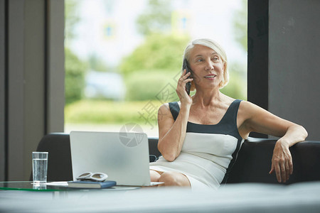 坐在沙发上在笔记本电脑前用手机交谈和在现代办公室微笑的成熟办图片