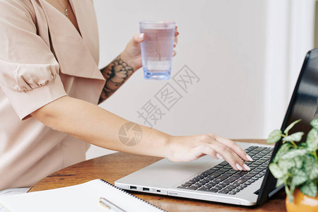 在笔记本电脑上工作检查电子邮件或观看网络研讨会的有淡水杯子的年轻女图片