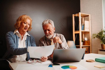 金发老妇人和灰色头发男子在办公桌旁看印刷图表时的近照图片