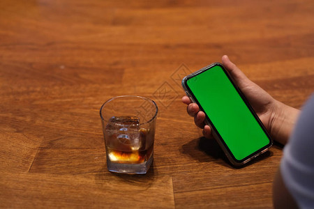使用绿色屏幕智能电话的人肩上在木桌上一杯冰饮料图片