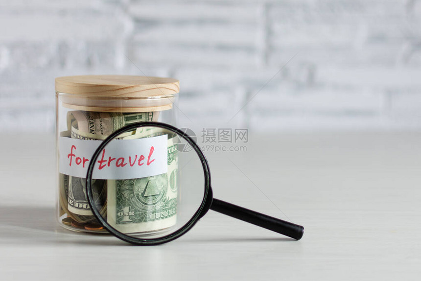 在白色砖墙上的白色木桌上用放大镜在玻璃罐中旅行的省钱旅行预图片