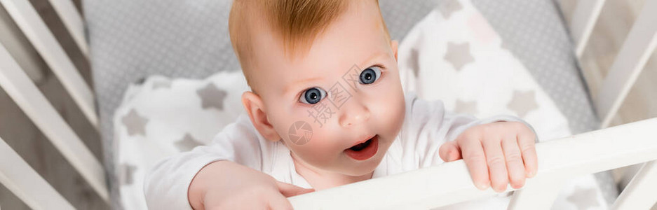 婴儿男孩站在婴儿床旁看照相机的横向概念图片