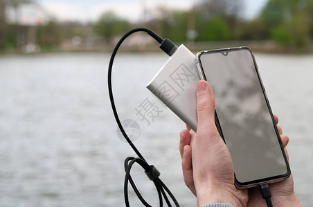 男子从河岸上的移动电源充电时使用带空白屏幕的智能手机现代技术概念图片