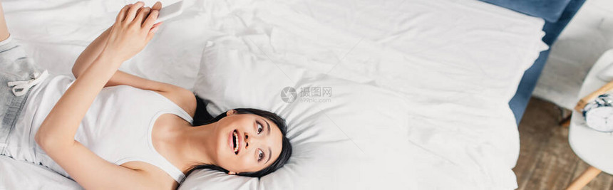 乐观的亚洲女孩持智能手机在床上视而不图片