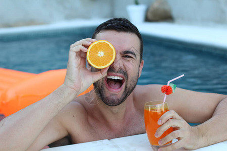 英俊的年轻男子在游泳池中将半个橙子放在眼图片