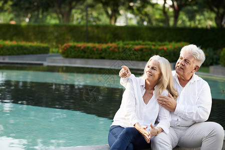 当他老婆坐在旅馆游泳池旁边时高年男子给妻子看一些有趣的东西图片