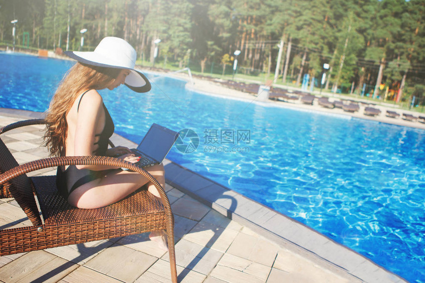 穿着黑色泳衣和沙滩帽的年轻美女在泳池附近用笔记本电脑工作一个追逐休息员图片