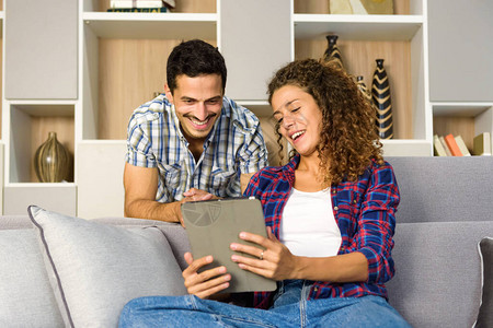 一对快乐的年轻夫妇在家里的客厅里一起看平板电脑上的媒体图片