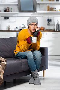 穿着热帽袜子和无指手套的冷冻男子在厨房里举茶杯时图片
