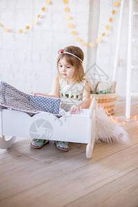 穿着白色裙子的可爱小女孩在明亮儿童房间的娃床上玩耍用玩具图片