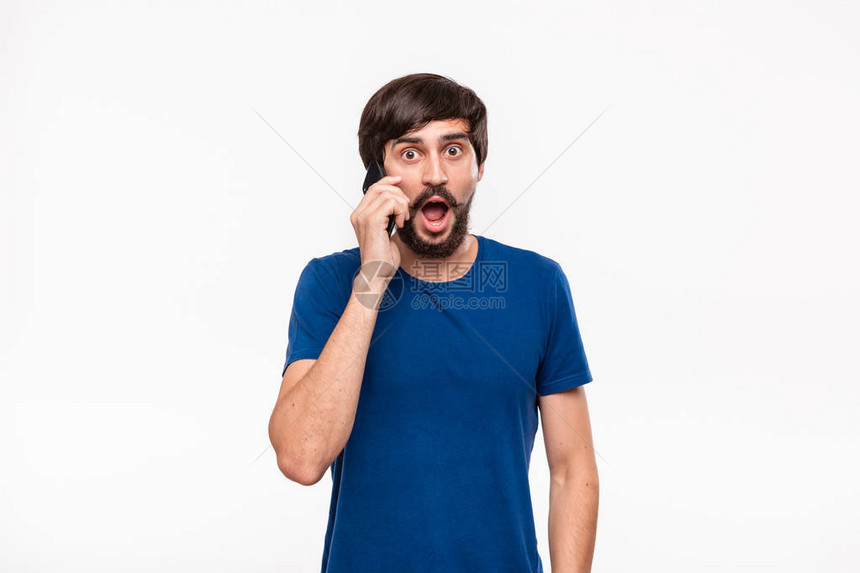穿着蓝色衬衫留着胡须和mostaches的漂亮深色男人震惊地拿着智能手机看着相机站在白色背景上孤立惊喜的情绪电话里传来意图片