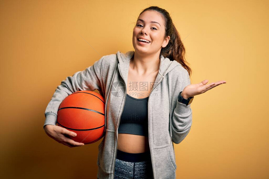年轻漂亮的褐发女运动员拿着篮球在黄色背景上非常高兴和奋图片