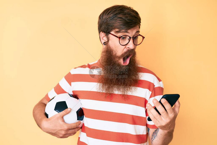 长胡子拿着橄榄球的帅气年轻红头男子看着智能手机庆祝疯狂和惊讶的成功图片