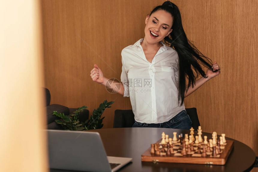 在棋盘和桌上笔记本电脑附近显示赢家姿态的兴奋女图片