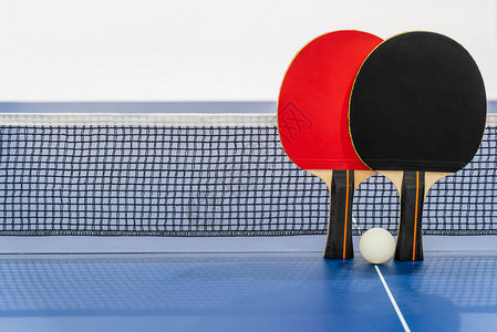 黑色和红色乒乓球拍和带网的蓝色乒乓球桌上的白球图片