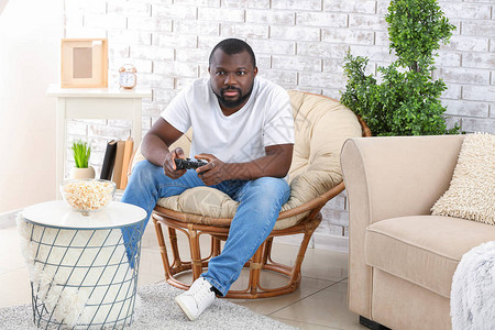 在家里坐在扶手椅上玩电子游戏的非裔美国人男子在图片