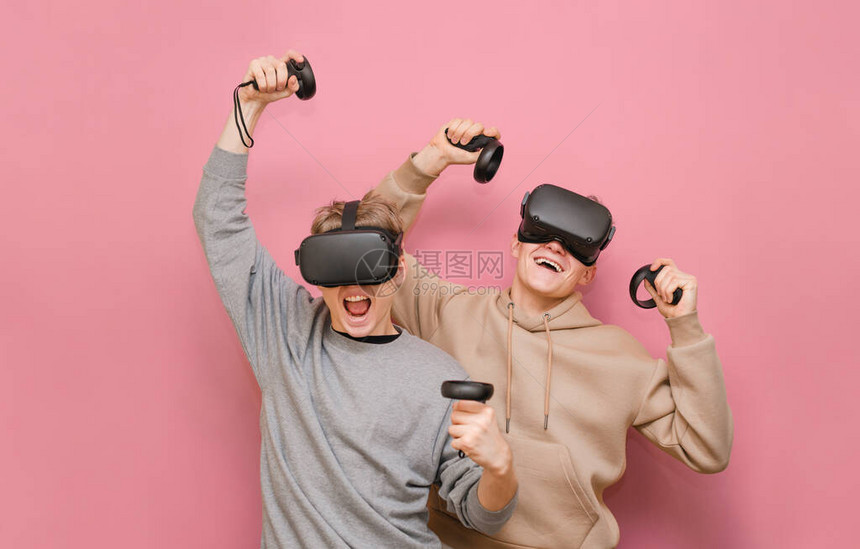 两个戴着VR头盔的快乐朋友享受着赢得比赛的乐趣2个人团队在粉红色背景下玩游戏孤立在虚拟现实中与朋友休闲图片