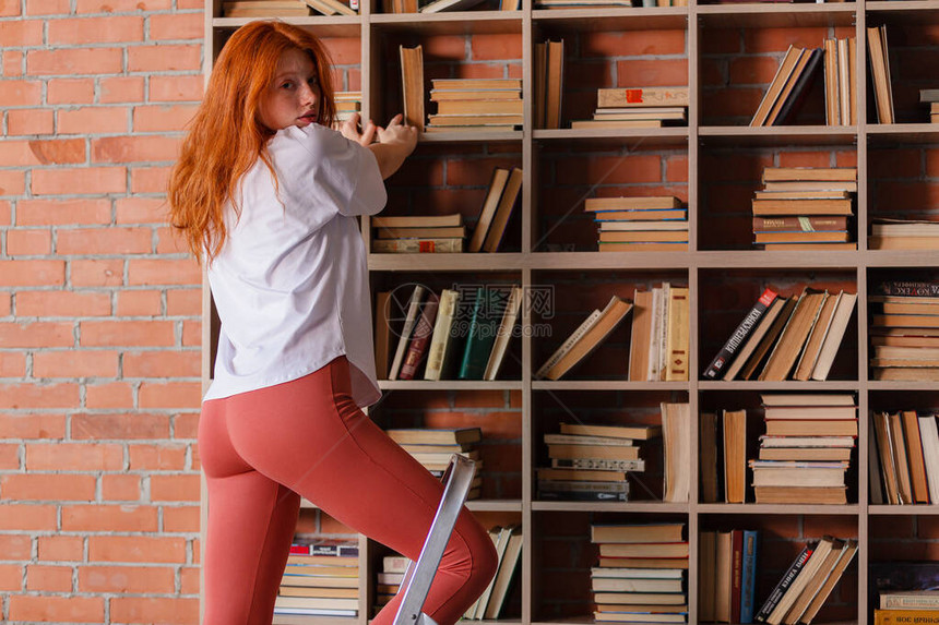 红发女大学生从图书馆的书架上拿书站在楼梯上从顶部拿一本书侧视图片