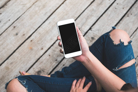 一名妇女坐在旧木地板背景上手持带空白黑屏的白色手机的顶图片