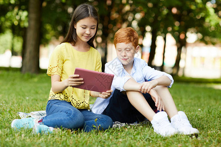 穿着黄色T恤坐在草地上向同学展示平板电脑上的节目图片