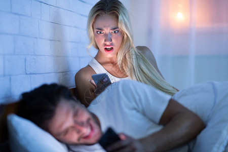 不忠嫉妒的女朋友抓到男朋友在智能手机上和其他躺在床上的女人一起发短信低光背景图片