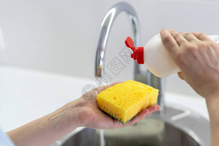 妇女洗碗器将肥皂倒在海绵上站在水池附近的白色明亮厨房的切图片