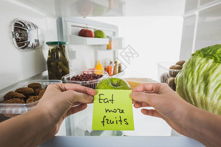 妇女持卡片多吃果实和在冰箱中以白色隔着的冰箱背景图片