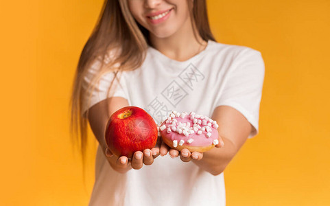 面目全非的微笑女孩拿着苹果和甜圈在镜头前提供健康或不健康的食物图片