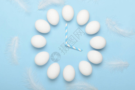 以挂钟形式的白色鸡蛋在蓝色背景与羽毛图片