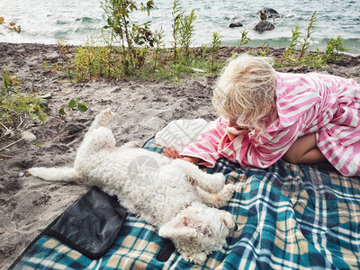 可爱的金发白种女孩抚摸躺在水湖河附近的海滩岸边的小白狗图片