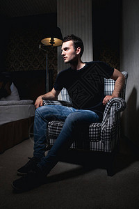 身穿黑色T恤的黑发强壮英俊男子坐在阴影中方格椅上的房间里图片