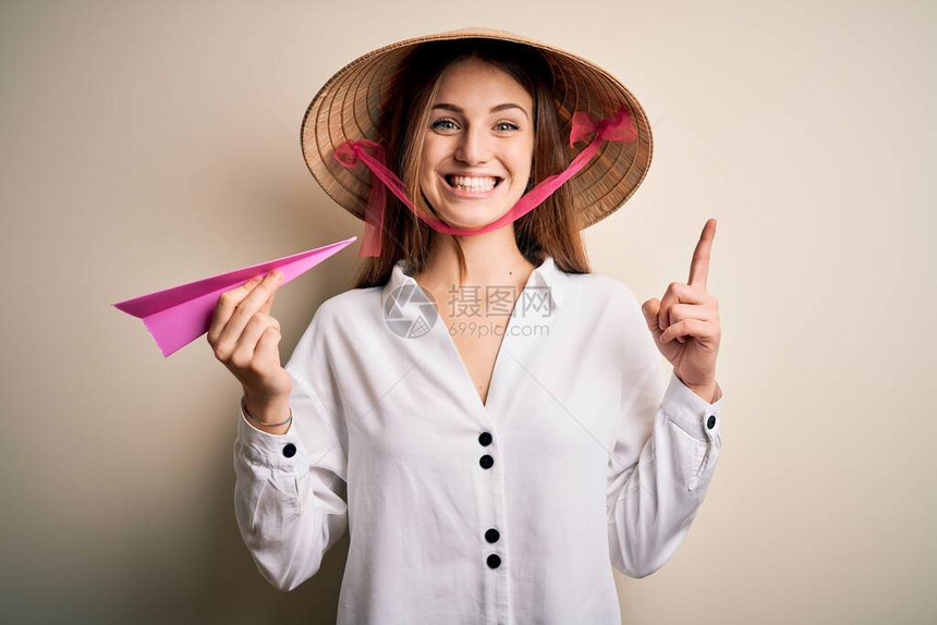 戴着亚洲传统帽子拿着纸飞机的年轻漂亮红发女人惊讶地发现一个想法或问题图片