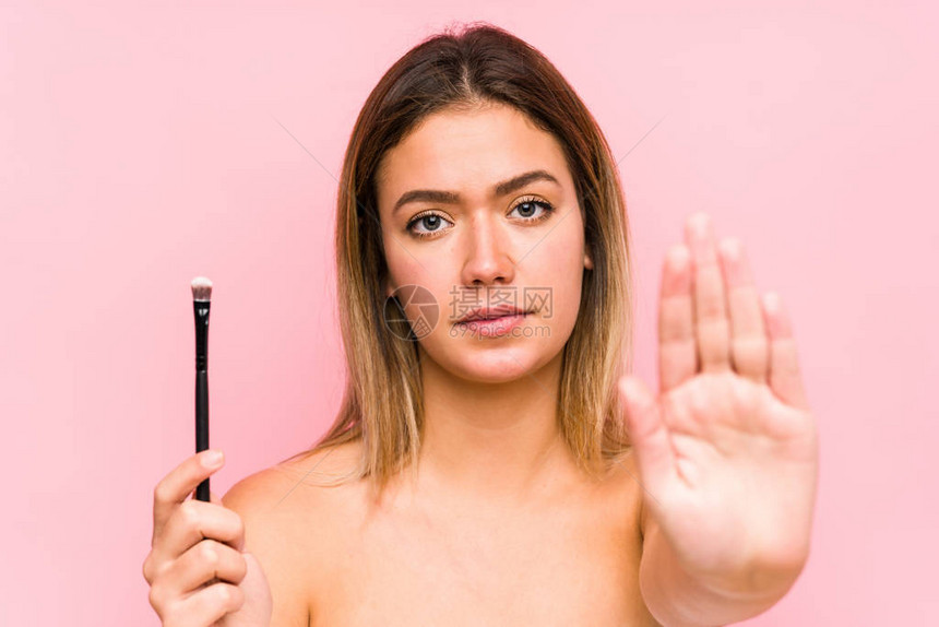 年轻的caucasian女人拿着一只孤立的眼笔站着伸展的手显示停图片
